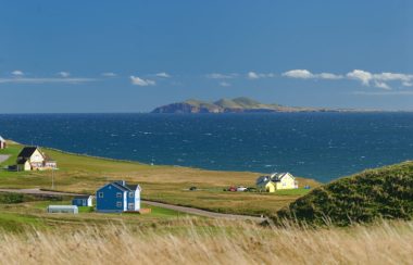 Trois maisons dans un champ en face de la mer avec une île en arrière plan.