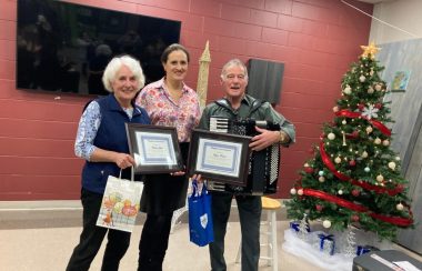 Deux femmes et un homme avec sacs de cadeaux et certificats en avant d'un Arbre de Noël décoré.