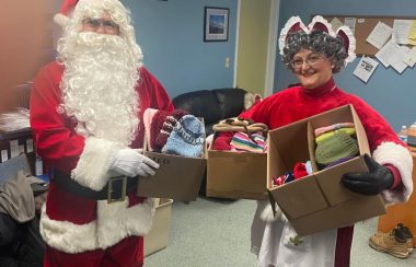 Homme déguisé en Père Noël et femme déguisée en Mère Noël tenant des boîtes de tuques multicolores.