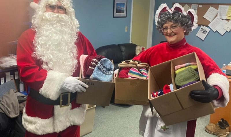 Homme déguisé en Père Noël et femme déguisée en Mère Noël tenant des boîtes de tuques multicolores.