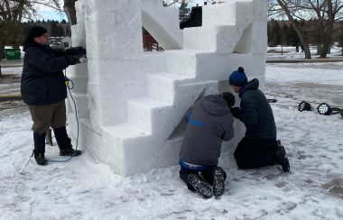 3 hommes qui travaillent sur une sculpture en neige. La sculpture présente une escalier qui monte de la gauche vers la droite. Une poutre de neige verticale se trouve vers le bas de l'escalier à la gauche. Les manteaux des hommes sont noir, gris et bleu.