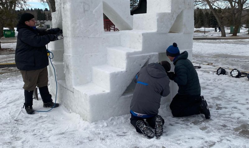 3 hommes qui travaillent sur une sculpture en neige. La sculpture présente une escalier qui monte de la gauche vers la droite. Une poutre de neige verticale se trouve vers le bas de l'escalier à la gauche. Les manteaux des hommes sont noir, gris et bleu.