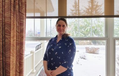 Pictured is executive director of Au Diapason Charlotte Evans standing in front of a window. She is wearing a navy blue floral shirt.