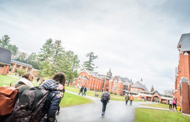 Students walking on a paved path in an open green space surrounded by historical brick buildings.