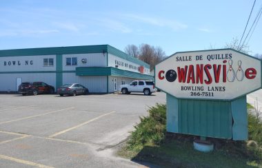 Pictured is the entrance to the Salle de Quilles Cowansville. Its building is grey with green accents. It's sign has Cowansville in bold red lettering with the O replaced by a bowling ball and the Ls by two bowling pins.