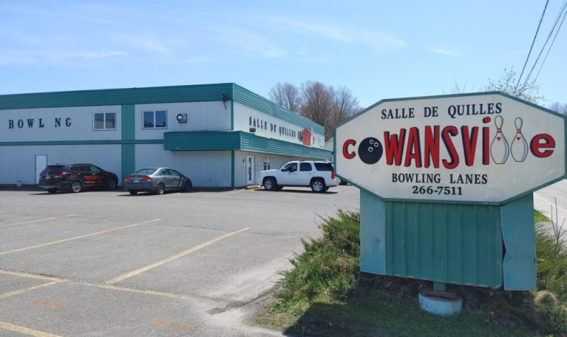 Pictured is the entrance to the Salle de Quilles Cowansville. Its building is grey with green accents. It's sign has Cowansville in bold red lettering with the O replaced by a bowling ball and the Ls by two bowling pins.