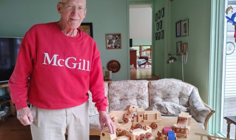 Pictured is Bob Pincott in his red McGill University sweater standing beside some of his wooden toys.