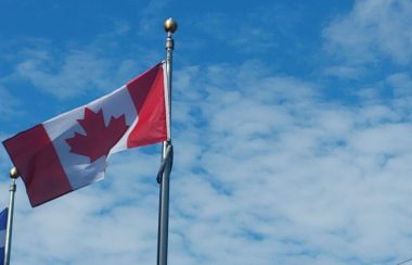 Pictured is the Canadian flag flying in the wind. It has to red stripes and one white stripe in the middle with a red maple leaf.