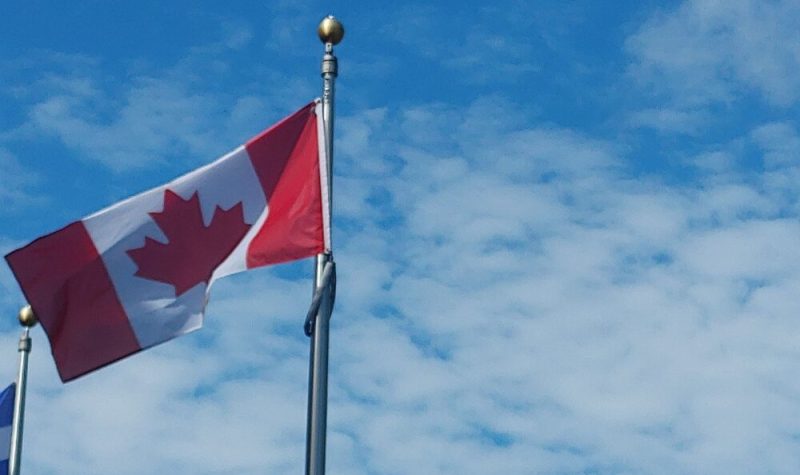 Pictured is the Canadian flag flying in the wind. It has to red stripes and one white stripe in the middle with a red maple leaf.