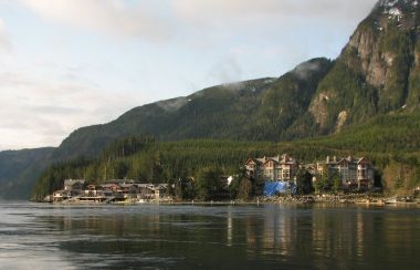 A series of hotel like buildings spread out across the shoreline, a heavily treed hillside rises up behind them