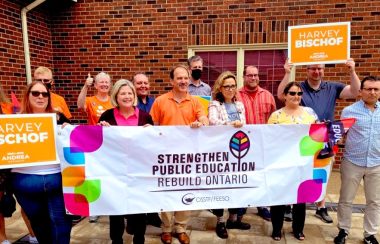 People in a row holding a large white sign with colours and text on it. Around the white sign are people holding up smaller orange signs.