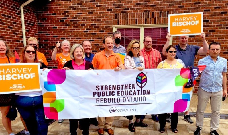 People in a row holding a large white sign with colours and text on it. Around the white sign are people holding up smaller orange signs.