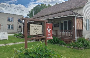 Pictured is a side shot of the Marché des Artisanats Dunham and its business sign. It is a cream coloured building with a sloping roof and a front porch. Inside it is two stories filled with arts and crafts.