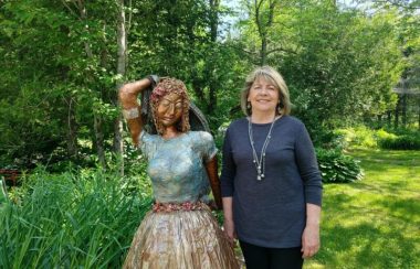 Pictured is sculptor Liane Chacra standing in her garden with one of her sculptures. The sculpture is a woman wearing a gold skirt, with a blue top, holding her arm up over her head.