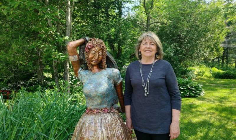 Pictured is sculptor Liane Chacra standing in her garden with one of her sculptures. The sculpture is a woman wearing a gold skirt, with a blue top, holding her arm up over her head.