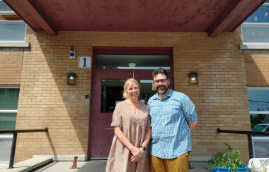 Catherine Canzani (left) and Nicholas Robert, standing in front Farnham Elementary School. It is a small brick building with a large red front door.