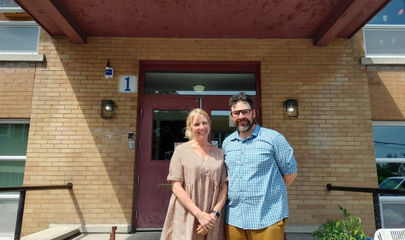 Catherine Canzani (left) and Nicholas Robert, standing in front Farnham Elementary School. It is a small brick building with a large red front door.