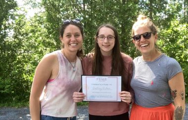 Jessica Adams and Jaime Moar awarding Destiny Picken, who is standing in between them, with the 2021 FEMS Award. They are standing outside with trees serving as the background.