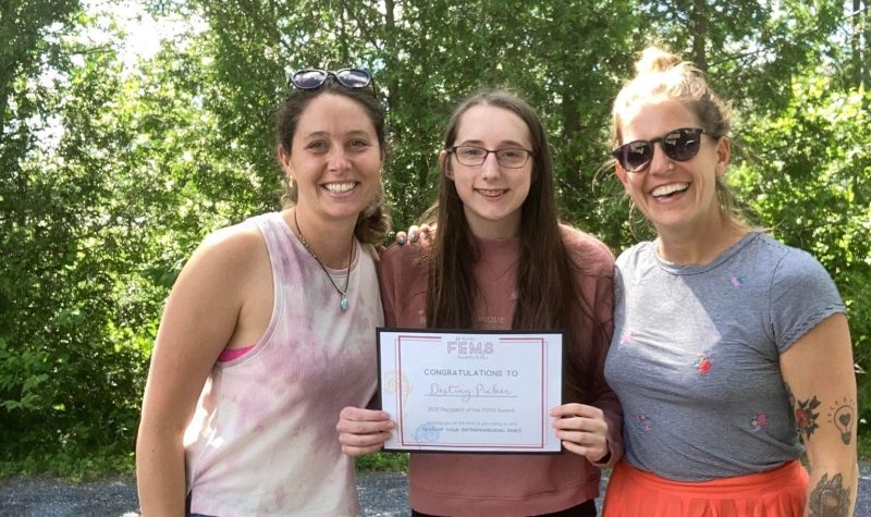 Jessica Adams and Jaime Moar awarding Destiny Picken, who is standing in between them, with the 2021 FEMS Award. They are standing outside with trees serving as the background.