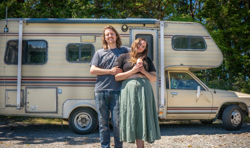 A man and a woman stand next to each other smiling in front of a camper van