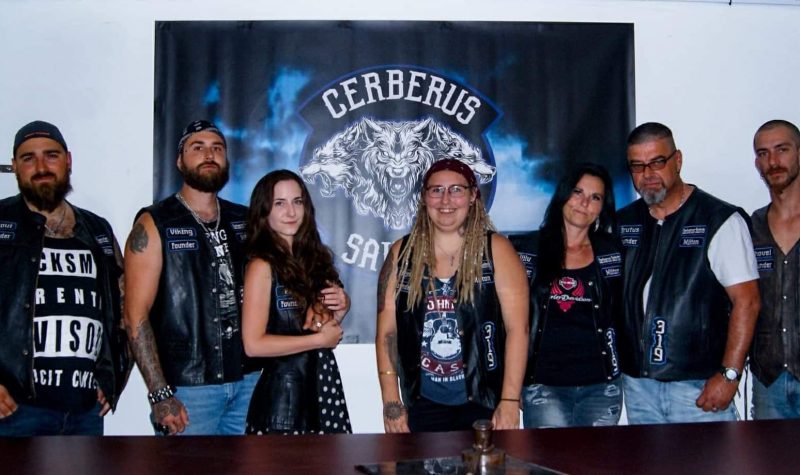 Pictured are members of the club dressed in their riding gear standing in front of a large printout of their patch. Their patch has a wolf in the centre, with dark blue colours, with the club name.