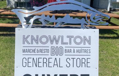 The business sign for Knowlton General. On the top of the sign there is a blue fish installation. The sign is in black and white and reads Knowlton on the top and General Store on the bottom. Between those two words is written Marché & Resto, Bio, Bar à Huîtres.