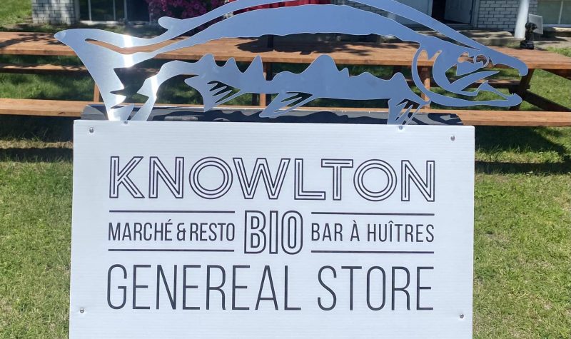 The business sign for Knowlton General. On the top of the sign there is a blue fish installation. The sign is in black and white and reads Knowlton on the top and General Store on the bottom. Between those two words is written Marché & Resto, Bio, Bar à Huîtres.