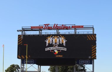 Large black scoreboard with red lettering at the top of it with. A design of stick people can be seen in the center of the scoreboard.
