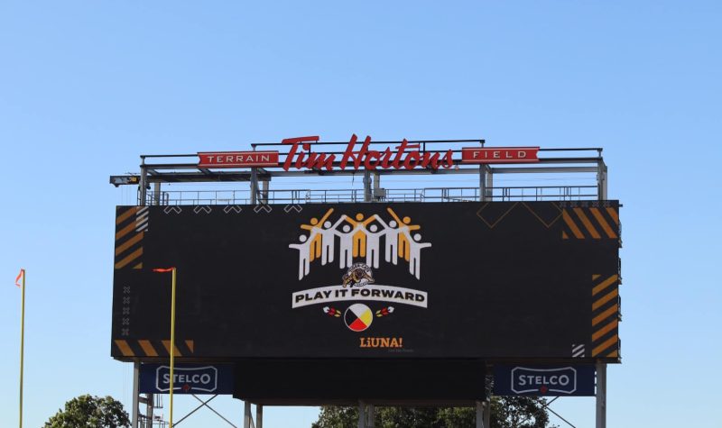 Large black scoreboard with red lettering at the top of it with. A design of stick people can be seen in the center of the scoreboard.