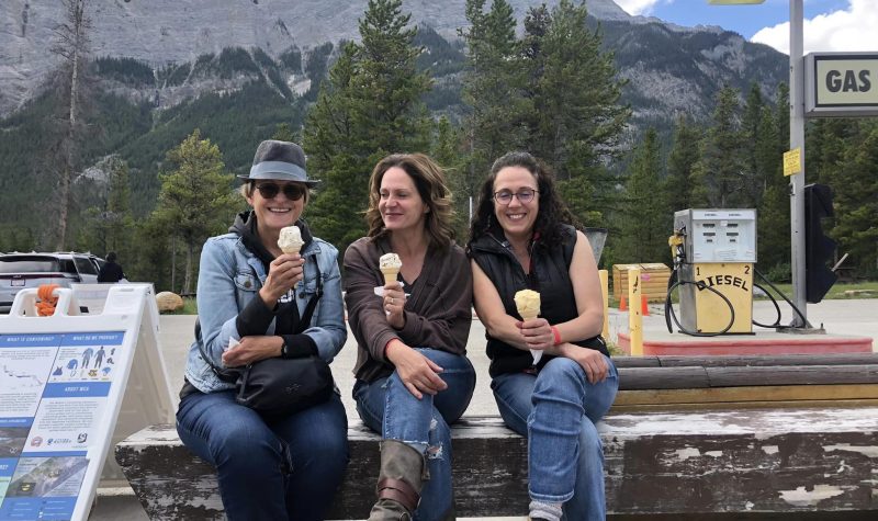 Trois dames sont assises sur un banc de bois foncé. Elles mangent de la crème glacée. Derrière elles, il y a une pompe à essence, des sapins et de grandes montagnes. Les trois dames sourient.