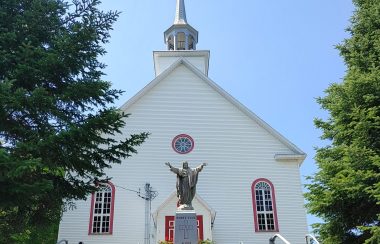 Pictured is the front of L’Église de Sainte-Étienne-de-Bolton. It is white with red borders around the windows. There is also a large statue of Jesus.