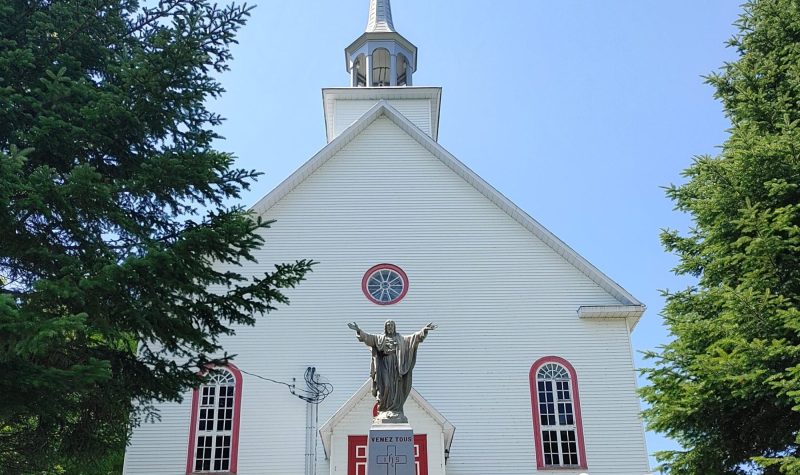 Pictured is the front of L’Église de Sainte-Étienne-de-Bolton. It is white with red borders around the windows. There is also a large statue of Jesus.