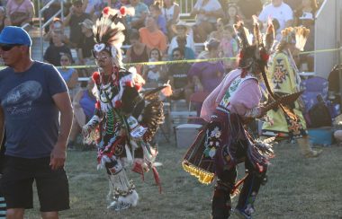 Traditional indigenous dancers can be seen performing in front of a grand stand filled with spectators. The dancers are wearing traditional indigenous regalia.