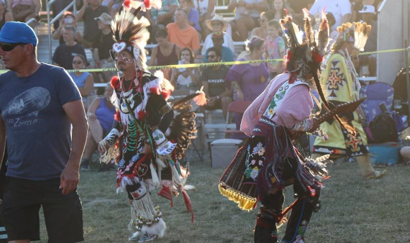 Traditional indigenous dancers can be seen performing in front of a grand stand filled with spectators. The dancers are wearing traditional indigenous regalia.