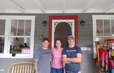 From left to right is Michel, Nancy, and Marc, standing in front of the check-in house at Les Minis. The building is grey with a red and white door.