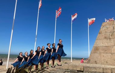 Des jeunes filles portant des blouses noires et des jupes bleues en avant de plusieurs drapeaux.