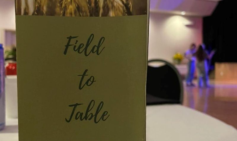 Le dépliant de l'événement est photographié de près, posé sur une table, avec des personnes discutant ensemble en arrière-plan.