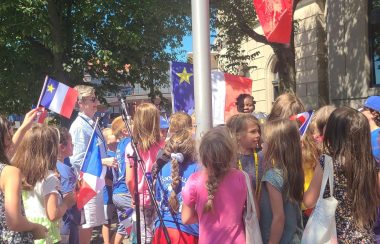 La mairesse Reardon est entourée d'enfants pour la levé du drapeau Acadien. Plusieurs enfants souriants sont visibles, ils portent des drapeaux acadiens.