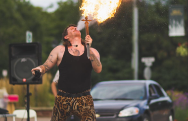 A man in a black tank top and animal print leggings breathes fire, on a street with sound system equipment surrounding him.