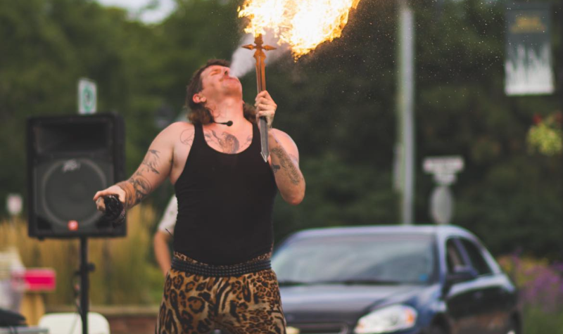 A man in a black tank top and animal print leggings breathes fire, on a street with sound system equipment surrounding him.