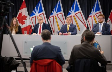Four people sit at a news conference table with the NL and Canada flags in the background.