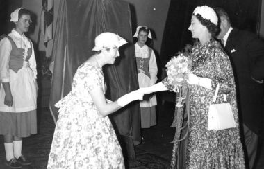 Une dame porytant un chsapeau, une longue robe et des gants donne la main à la Reine.