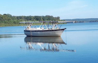 Un voilier, sans mat et sans voiles, flotte sur l'eau, avec une forêt en arière-plan.