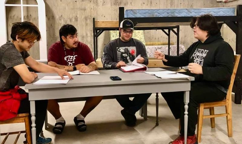 Four male cast members rehearse a scene around a table.