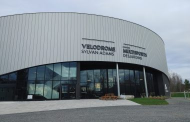 Pictured is the front of the velodrome. The top half of the velodrome is grey with the bottom half being made up of windows and doors. ylvan Adams Velodrome Multi-sport Centre Desjardins, the name of the velodrome, can also be seen on the front of the building.