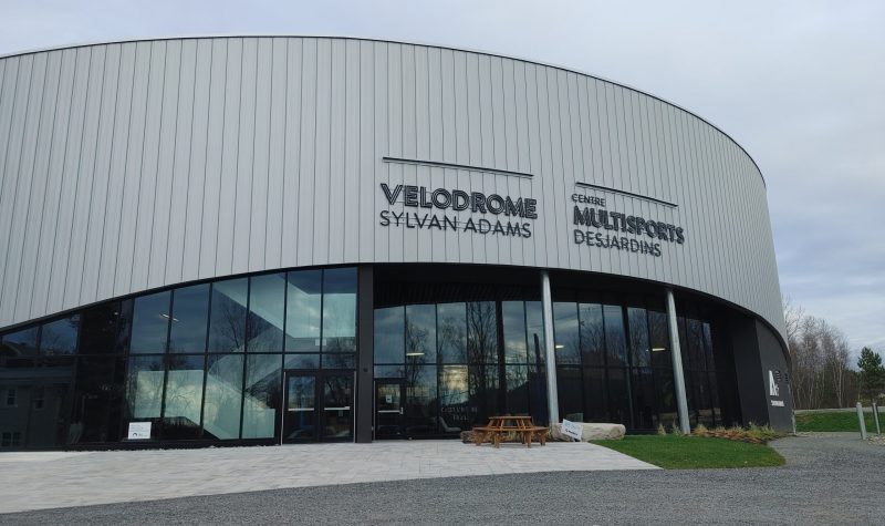 Pictured is the front of the velodrome. The top half of the velodrome is grey with the bottom half being made up of windows and doors. ylvan Adams Velodrome Multi-sport Centre Desjardins, the name of the velodrome, can also be seen on the front of the building.