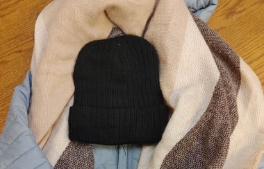 A light blue jacket layered with a scarf and a hat. There is a wood floor in the background.