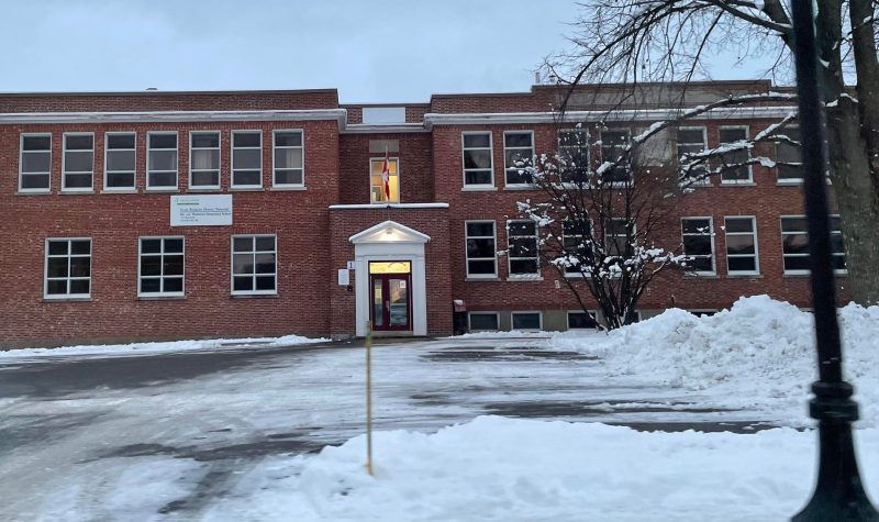 The front of a two story elementary school. It is a brick building with large windows that have white trim. There is snow in front of the school.
