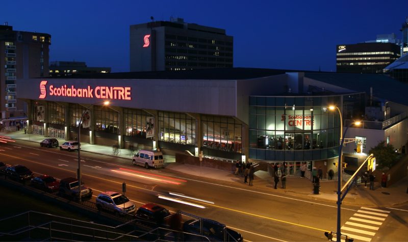 Landscape photo taken from across the street of the Scotiabank Centre. People stand outside of the venue with streetlights lighting the street. Scotiabank Centre sign lit up in red.