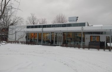 Pictured is the front of Theatre Lac-Brome building. The front is made up of large glass windows and the building itself is rectangular. The building is white and grey.